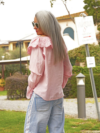 Ältere Frau trägt die OFF#DLY Bluse Greta rose mit Spitzenkragen und weiten Ärmeln, kombiniert mit lässigen Jeans, in einem Garten stehend, verkörpert zeitlose Eleganz mit romantischen Details.