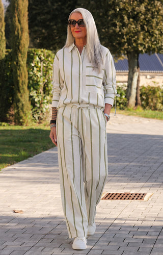 Frau in Co Couture Idie Stripe Pant offwhite/green spaziert auf einem Gartenweg, trägt ein locker sitzendes, gestreiftes Set mit Sneakern, zeigt Armbanduhren und ein Tattoo auf dem Unterarm.