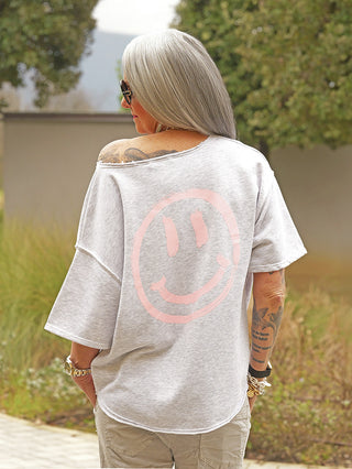 Person mit langen grauen Haaren trägt den OFF#DLY Sweater Smiley grey mel. Der Sweater hat einen großen Smiley-Print auf dem Rücken und eine lässige Off-Shoulder-Optik.