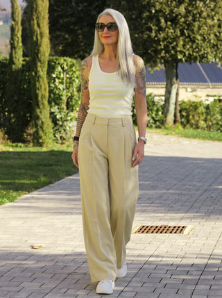 Die Co Couture Nilan Pleat Pant zeigt sich in einem eleganten, weiten Schnitt, der eine moderne Silhouette formt. Sie ist kombiniert mit einem gestreiften Tanktop und weißen Sneakern.