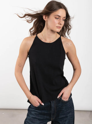 Junge Frau trägt OUTRYGHT RIB Top Tina faded black, ein schwarzes, geripptes Halter-Top, locker in dunkle Jeans gesteckt, mit goldenen Accessoires und sichtbaren Tattoos auf den Armen.