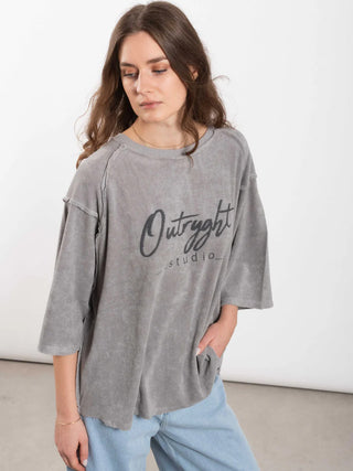 Eine junge Frau trägt das Outryght Loo T grey moon, ein locker sitzendes Pullover-Oberteil in gewaschenem Grau, kombiniert mit hellblauen Jeans, minimalem Schmuck und natürlichem Make-up.