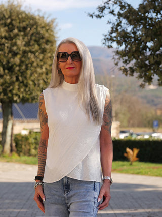Frau mit grauen Haaren trägt das Co Couture Conny Drape Top in Weiß, mit asymmetrischer Drapierung und hochgeschlossenem Kragen, kombiniert mit Jeans und Accessoires, auf einem sonnigen Pfad.