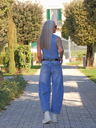 Person in OFF#DLY Barrel Denim Ina midblue Jeans, mit weitem Barrel-Fit, läuft auf Gartenweg. Die Jeans zeigt eine lässige Silhouette, kombiniert mit weißen Boots und einem ärmellosen Denim-Oberteil.