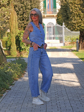 Ältere Frau in OFF#DLY Barrel Denim Ina midblue; modische Barrel-Silhouette, ärmellose Jeansweste, weiße Boots, Tattoos am Arm, Sonnenbrille, Accessoires, sonnige Gartenumgebung.