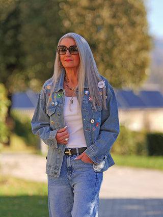 Eine Frau trägt die OFF#DLY Denimjacke Patch midblue mit zahlreichen Patches und Pins, kombiniert mit einer Jeans. Die Jacke hat einen lässigen Schnitt und Brusttaschen.