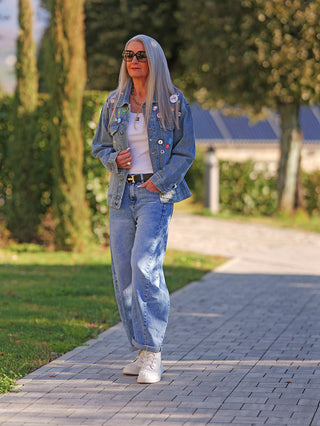 Eine Frau trägt die OFF#DLY Denimjacke Patch midblue, verziert mit kleinen Pins, über einem weißen Tanktop. Die Jacke hat einen lässigen Schnitt und viele Statement-Patches.