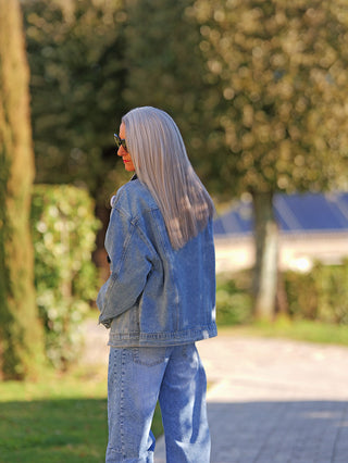 Eine Person trägt die OFF#DLY Denimjacke Patch midblue mit auffälligen Patches und entspanntem Schnitt. Sie kombiniert die Jacke lässig mit passenden Jeans in einer sonnigen Outdoor-Szene.