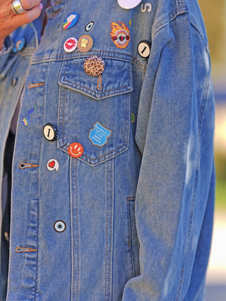 Die OFF#DLY Denimjacke Patch midblue zeigt eine Vielzahl kleiner Pins, darunter ein Leoparden-Button und ein Stronger Than Ever-Flammenmotiv, die dem klassischen Design eine verspielte Note verleihen.