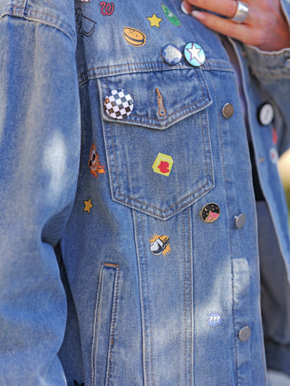 Nahaufnahme einer getragenen OFF#DLY Denimjacke Patch midblue, verziert mit diversen bunten Pins und Patches, darunter ein schwarz-weißes Karomuster, ein Stern, ein Cheeseburger-Icon und ein KXLE-Abzeichen.