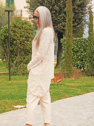 Ein Erwachsener trägt das OFF#DLY Longshirt Spitze Tina beige, ein elegantes Longshirt mit fließendem Schnitt und strukturierter Spitze, während er draußen auf einem Weg in einem Garten steht.