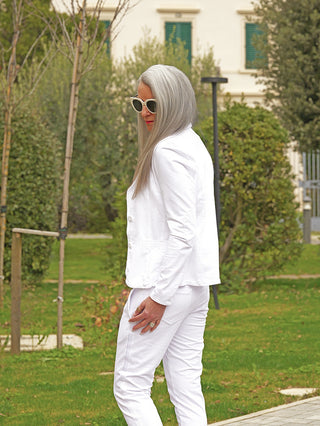 Person im Garten mit dem OFF#DLY Blazer Britt white, eine schmal geschnittene, elegante Jacke aus Sweatmaterial, kombiniert mit weißer Hose und weißen Sonnenbrillen, vor einem Gebäude mit geschlossenen Fensterläden.
