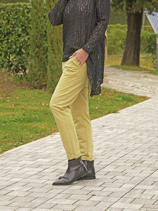 Eine Person trägt die OFF#DLY Pant Cleo matcha, eine schmal geschnittene Hose in modernem Stil, kombiniert mit einem langen, schwarzen Spitzen-Tunika-Top und schwarzen Lederstiefeletten.