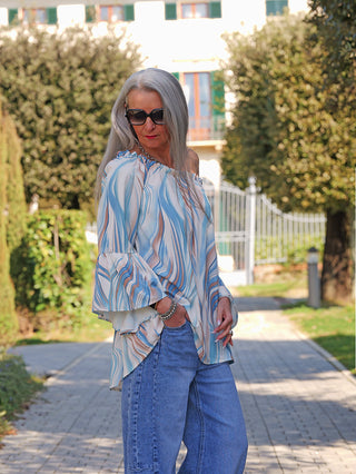 Frau trägt das OFF#DLY Blusenshirt Biggi blue multi mit lockerer Passform und Volantsärmeln, auf einem Gartenweg stehend, kombiniert mit blauer Jeans, Accessoires und Sonnenbrille.