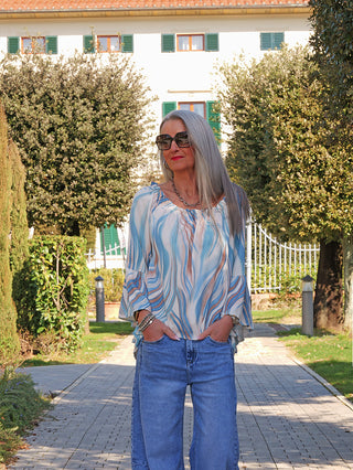 Frau im Garten trägt das OFF#DLY Blusenshirt Biggi blue multi, locker fallend mit voluminösen Ärmeln und eleganten Mustern, in Blau- und Naturtönen, passend zu Jeans.