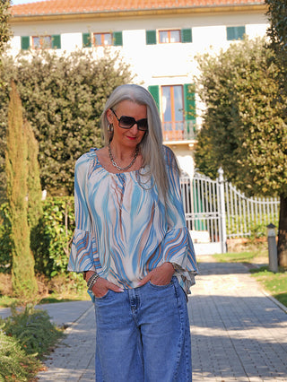 Die Frau trägt das OFF#DLY Blusenshirt Biggi blue multi mit fließendem, locker fallendem Design und voluminösen Volantärmeln, kombiniert mit weit geschnittenen Jeans, vor einem eleganten Hintergrund.