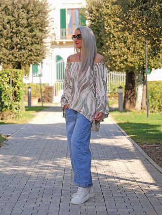 Eine Person trägt das OFF#DLY Blusenshirt Biggi green multi, mit lockerer Passform und verspielten Volants, während sie auf einem gepflasterten Gartenweg posiert.