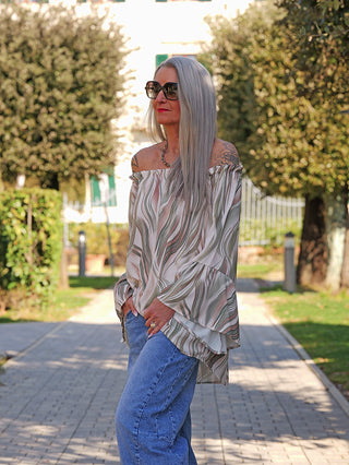 Erwachsene Frau trägt das OFF#DLY Blusenshirt Biggi green multi mit fließendem, schulterfreiem Design und Volantärmeln, kombiniert mit lässigen Jeans, auf einem Gartenweg.