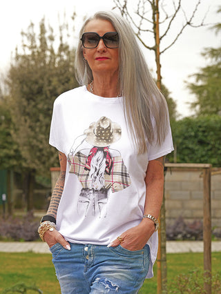 Frau in weißem OFF#DLY Tee Hat red mit Cowboy-Illustration, oversized Schnitt, kombiniert mit hellen Jeans, silbernem Schmuck und Tätowierung im Park.