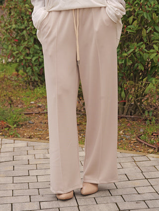 Person trägt die OFF#DLY Pant Palermo beige, eine locker geschnittene, fließende Hose mit elastischem Bund und Kordelzug, kombiniert mit passenden Slip-On-Schuhen auf einem Steinweg.