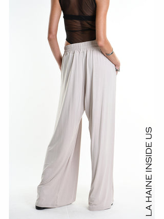 La Haine Pant 5271 beige aus Bamboo-Jersey mit weitem Palazzo-Schnitt, feinen Falten und elastischem Bund, getragen von einem Modell mit silbernen Armbändern, betont moderne Eleganz und Komfort.
