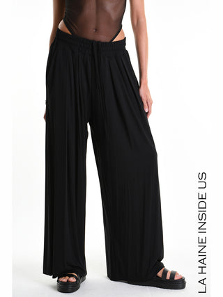 La Haine Pant 5271 schwarz: Weibliches Modell trägt fließende, schwarze Palazzo-Hose mit weitem Bein und elastischem Kordelzugbund, kombiniert mit schwarzen Plateau-Sandalen, Branding LA HAINE INSIDE US sichtbar rechts.