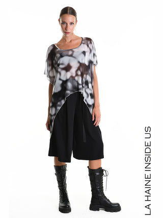 Modell trägt La Haine Tee 5349 white, ein asymmetrisches, locker geschnittenes Shirt aus bedrucktem Tüll, kombiniert mit schwarzen Culotte-Shorts und hohen Schnürstiefeln.