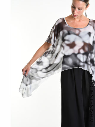 Frau trägt La Haine Tee 5349 white, ein asymmetrisches, semi-transparentes Oversize-Shirt mit abstraktem Muster, weitem Rundhals und fließendem Schnitt über weiten, schwarzen Hosen, zeigt das Shirt's Drapierung.