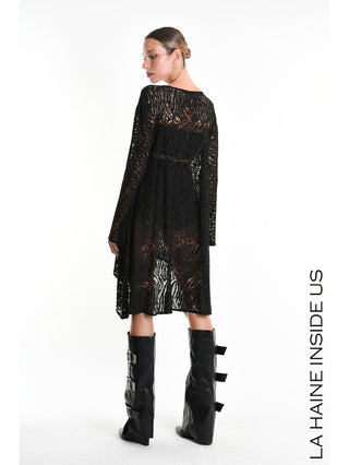 Model trägt das La Haine Dress 5377 black, ein knielanges, langärmeliges schwarzes Spitzenkleid mit asymmetrischem Saum. Kombiniert mit hohen, geschnallten Lederstiefeln, verkörpert es avantgardistische Eleganz.