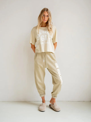 Person trägt Penn&Ink Tee Print oyster/white, ein lässiges, locker geschnittenes T-Shirt mit dem aufgedruckten Logo „SOHO CHAMPS 61 NEW YORK“, kombiniert mit beigen Jogginghosen und passenden Hausschuhen.