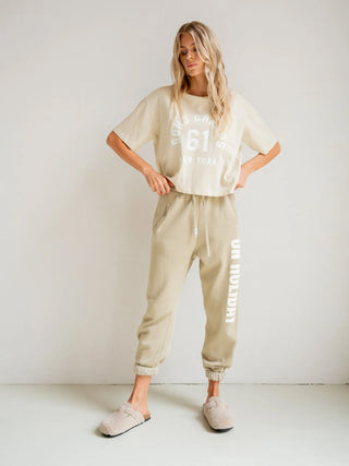 Penn&Ink Hose Print perrer/ecru: Frau trägt entspannte Jogpant mit „ON HOLIDAY“-Print, elastischem Bund und schmal zulaufender Silhouette, kombiniert mit lockerem Waffle-Knit-Top und neutralen Mule-Hausschuhen.