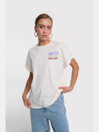 Eine junge Frau trägt das ALIX Club Tee cream mit einem kleinen ALIX CLUB – THE LABEL Print auf der Brust, kombiniert mit hellen Jeans und großen goldenen Ohrringen.