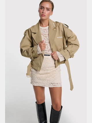 Junge Frau in einem ALIX Lace Top cream, einem figurbetonten, floralen Spitzenoberteil mit langen, leicht ausgestellten Ärmeln, kombiniert mit einem khaki-tan Trenchcoat und glänzenden schwarzen Stiefeln.