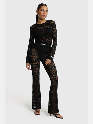 Modell trägt ALIX Lace Top black: figurbetontes, florales Spitzentop mit langen, leicht ausgestellten Ärmeln und Rundhalsausschnitt, kombiniert mit passenden, ausgestellten Spitzenhosen und schlichten, schwarzen Sandalen.