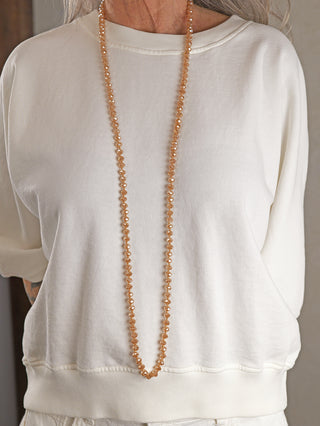Oberkörperansicht mit einer Glasperlenkette oval 7 mm beige in sanftem Pfirsichton, die über einem cremefarbenen Pullover getragen wird. Die lange Halskette aus facettierten Glasperlen ergänzt den schlichten Look.