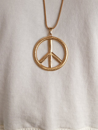OFF#DLY Peace Kette gold: Goldener PEACE-Anhänger an passender Textilkordel, präsentiert auf einem hellen, strukturierten Stoff. Durchmesser: 5,5 cm, Länge inkl. Anhänger 47 cm plus 5 cm Verlängerung.