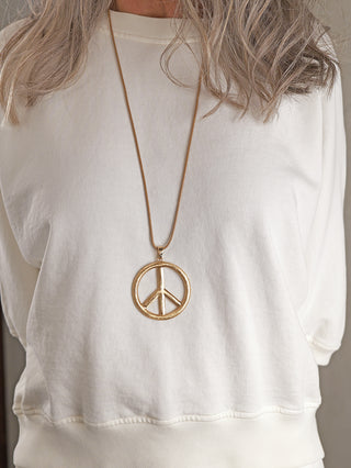Person trägt OFF#DLY Peace Kette gold mit großem, rundem PEACE-Anhänger an langem, dünnem Kettentextil. Das Gesicht ist nicht sichtbar, nur Oberkörper und Haaransatz.
