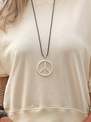 Nahaufnahme eines Torsos mit der OFF#DLY Peace Kette silber, einem großen, runden PEACE-Anhänger an einem dünnen, dunklen Textilband. Sichtbar sind ein tätowierter Unterarm und ein Perlenarmband.