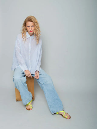 Person trägt Emily Bluse 157910 stripe white blue, eine locker sitzende, semi-transparente Hemdbluse mit zartem Blau/Weiß-Streifenmuster. Kombiniert mit weit geschnittenen Jeans und neon-gelbgrünen Sandalen.