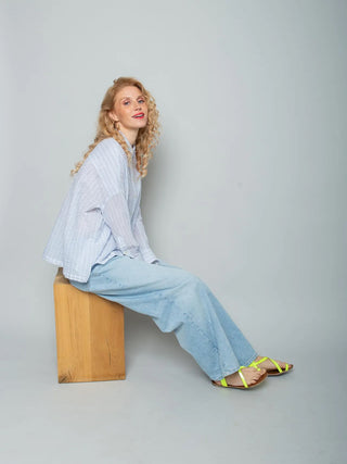 Junge Frau trägt Emily Bluse 157910 stripe white blue, eine lässige, durchgeknöpfte Hemdbluse mit zartem Blau/Weiß-Streifen und XL-Saumweit, kombiniert mit locker sitzenden Jeans und neon-gelben Sandalen.
