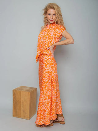 Emily Bluse 155690 orange/leo: Frau in seitlicher Ansicht trägt ein kurzes, gebundenes, leuchtend orange-pinkes Leo-Print-Oberteil aus Viskose, kombiniert mit passendem Rock und braunen Sandalen.