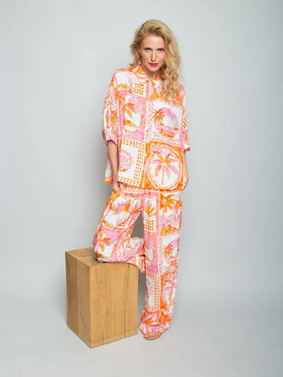 Emily Damenbluse 156844 orange/pink: Bluse mit Halbarm und Hemdblusenkragen, stilisiertem Scarf-Print aus Palmen und Motiven, getragen von einem Modell mit passender weiten Hose, Hände in den Taschen.