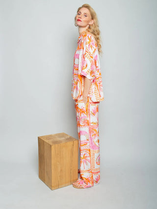 Emily Damenbluse 156844 orange/pink: Frau im Profil mit locker geschnittener, durchgeknöpfter Bluse in tropischem Print, kombiniert mit weiten Hosen. Bluse hat Hemdblusenkragen und schmale Manschetten.