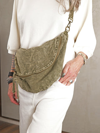 Person trägt OFF#DLY Crossbody Denim green, eine olivgrüne, gewaschene Canvas-Tasche mit verstellbarem Riemen und Zipper-Tasche. Hände mit metallic Nagellack, Ringen, Armbändern und Uhr sichtbar.