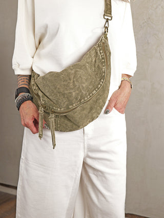 Person trägt eine OFF#DLY Crossbody Denim green, eine olivgrüne Sling-Tasche aus 100% Baumwolle mit doppeltem Reißverschluss vorne und verstellbarem Gurt, kombiniert mit weißer Kleidung und Armbändern.