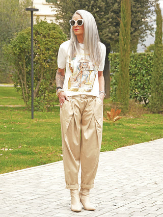 Schlanke Frau im Garten trägt OFF#DLY Hose Ida beige: High-waisted, Bundfalten, verkürzte Beine. Kombiniert mit weißem T-Shirt und Stiefeln, vermittelt lässige Eleganz und Komfort im modernen Schnitt.