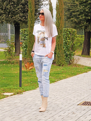 Frau in OFF#DLYDenim Destroy Camo lightblue Jeans mit Destroyed-Details und Camo-Patches, kombiniert mit weißem Grafik-T-Shirt und beigen Stiefeletten, steht lässig auf einem Gartenweg.