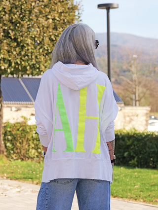Person von hinten in weißem OFF#DLY Hoody YEAH mit großen neonfarbenen Buchstaben, grauem Haar, Sonnenbrille, Jeans, auf Gehweg in parkähnlichem Umfeld.