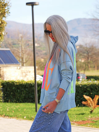 Person in einem Park trägt OFF#DLY Hoody YEAH lightblue mit auffälligem Neon-Print, lockerer Passform und Kapuze, kombiniert mit lässigen Jeans. Ideal für einen modernen, sportlichen Look.