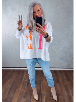 Person mit Smartphone in einem weißen Sweatshirt und OFF#DLY Denim Milano lightblue Jeans, locker und modern mit Destroy-Look, kombiniert mit spitzen Stiefeletten, steht vor einem Spiegel und zeigt das Peace-Zeichen.